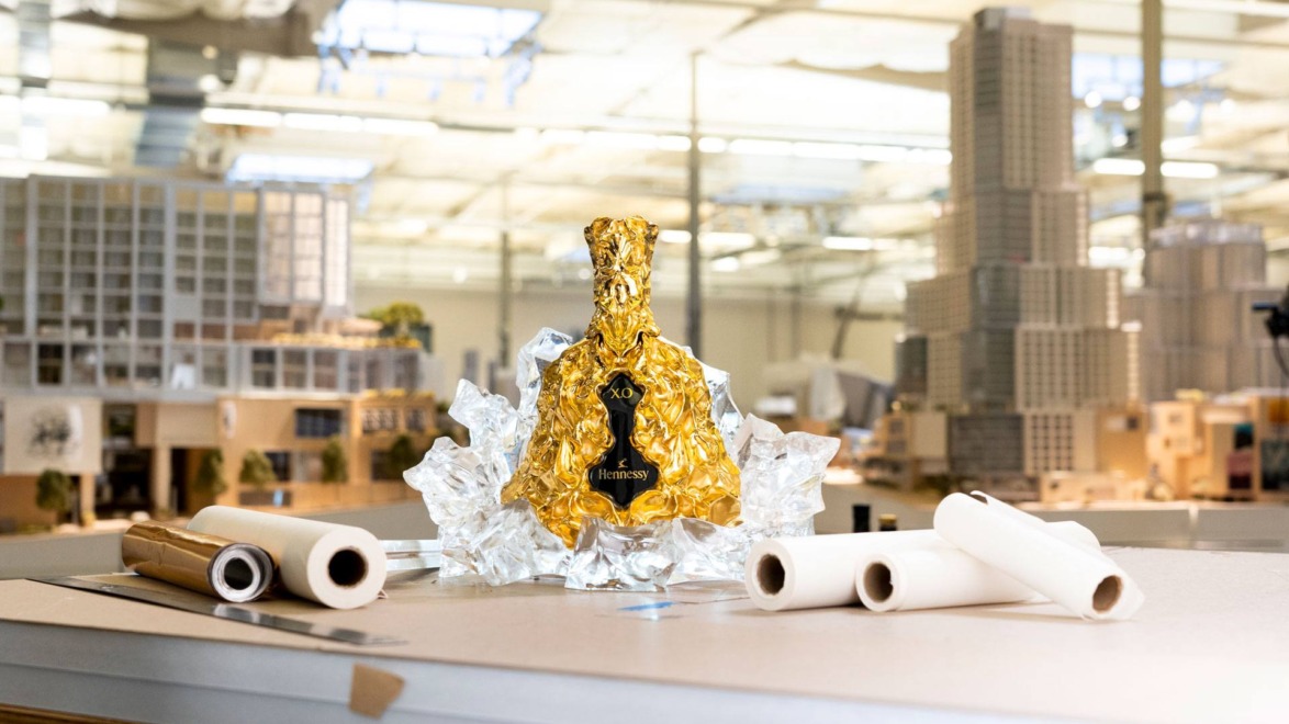 Golden bottle wrapped in foil, surrounded by ice, on a table with architectural models and rolled papers in the background.