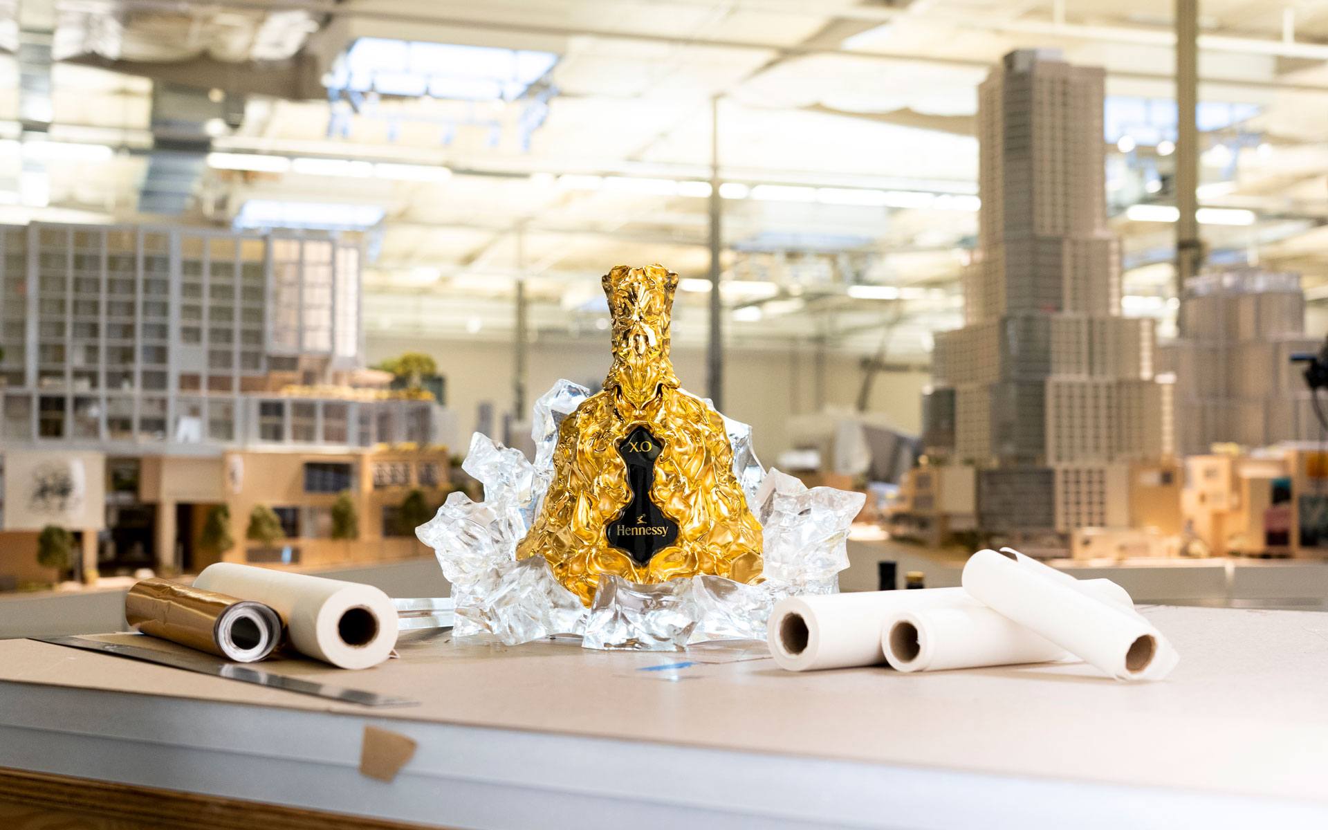 Golden bottle wrapped in foil, surrounded by ice, on a table with architectural models and rolled papers in the background.