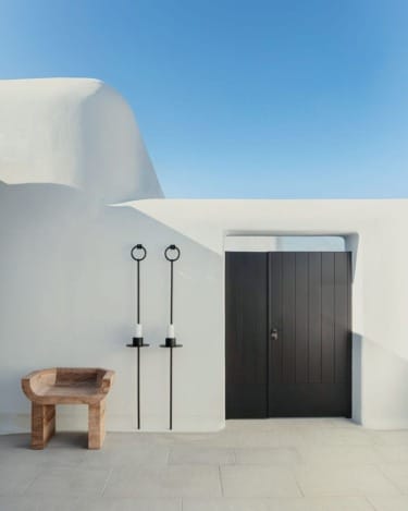 Minimalist white stucco building with black door, wooden chair, and two modern candle holders under a clear blue sky.