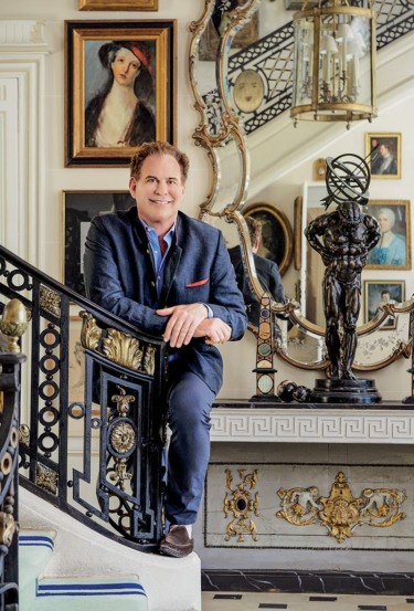 Man in a blue suit leaning on an ornate staircase railing, with mirrors and artwork in the background.
