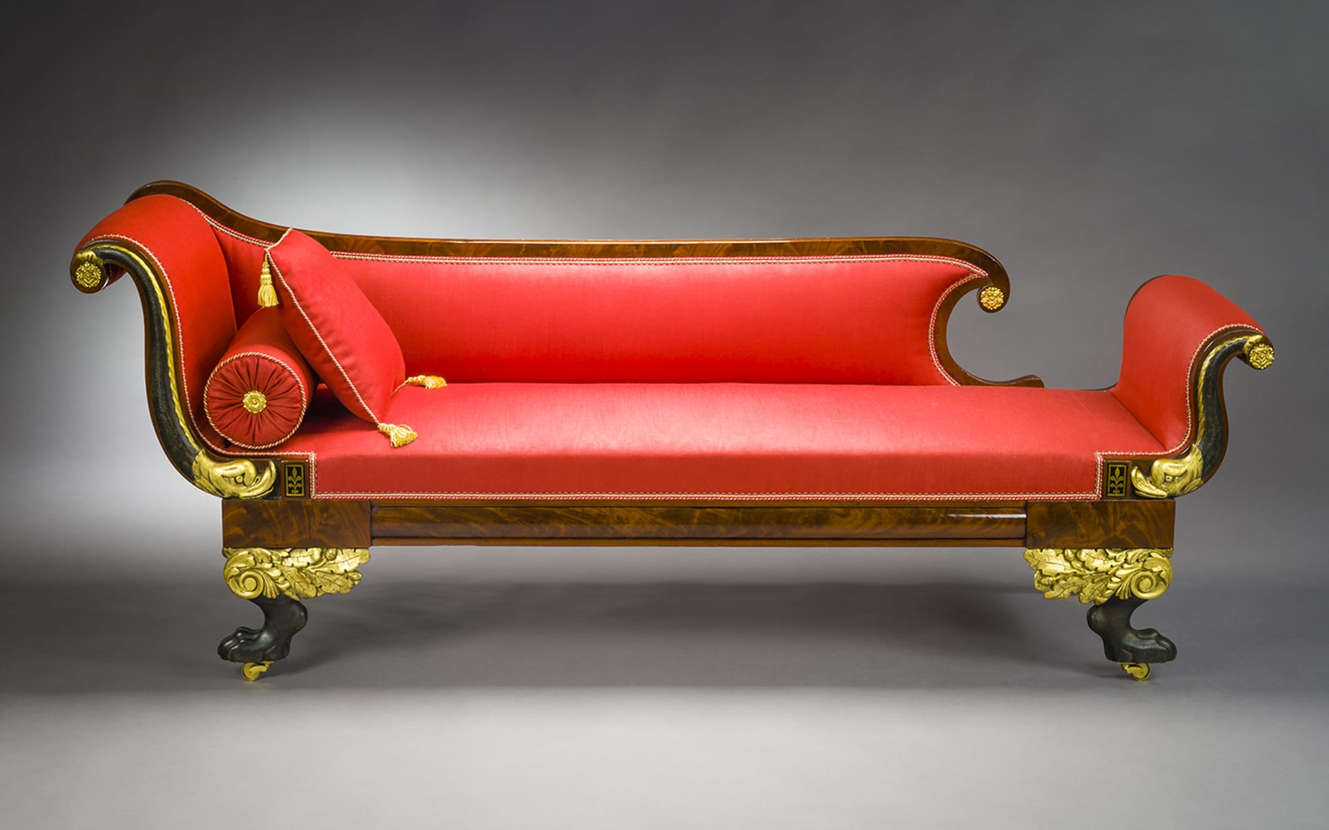 Red and gold ornate antique sofa with decorative cushions on a neutral background.