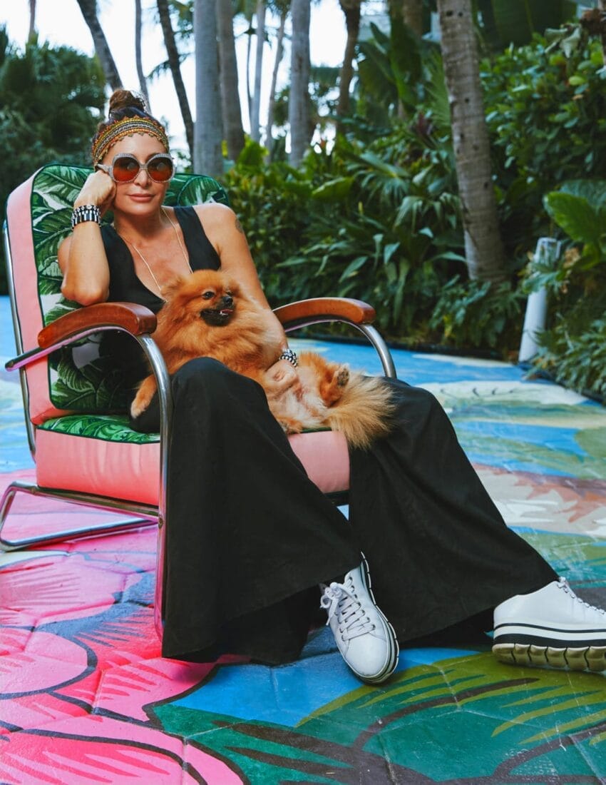 Person sitting on a colorful outdoor chair, holding a small fluffy dog, surrounded by lush greenery.