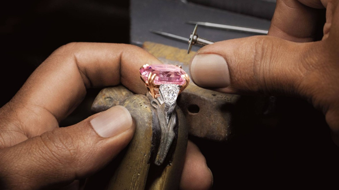 Craftsman adjusting a ring with a large pink gemstone and silver accents, using specialized tools on a wooden holder.