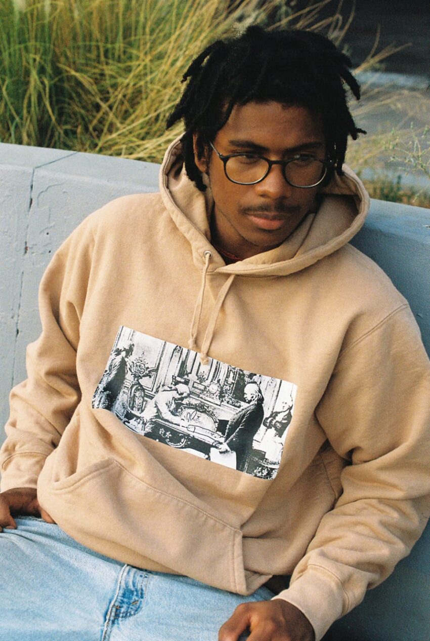 Person wearing glasses and a tan hoodie with a black and white design, sitting outdoors on a concrete bench.