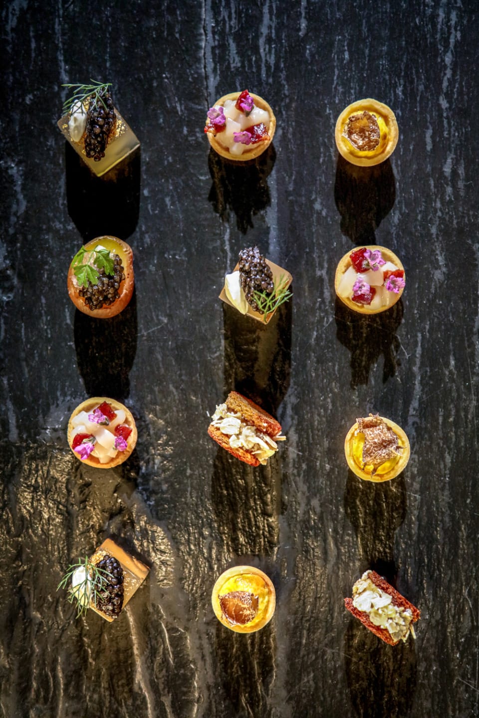Assorted gourmet appetizers on a dark slate surface, featuring caviar, herbs, and colorful garnishes arranged neatly.