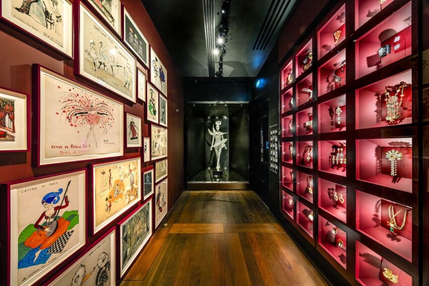 Art exhibit hall displaying framed sketches and jewelry in red-lit cases, with a sculpture at the end of the wooden-floored corridor.