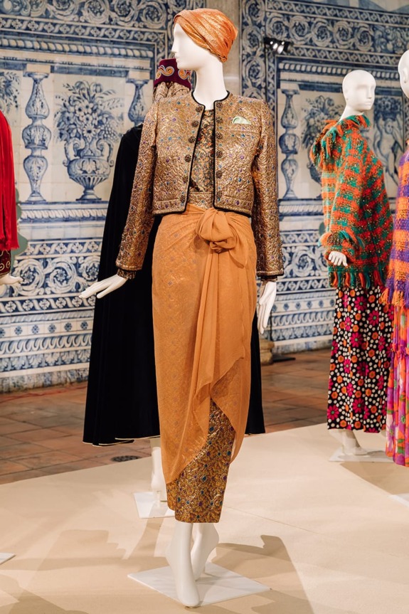 Mannequin wearing an ornate jacket with a long orange skirt, displayed in a room with detailed blue and white tiles.