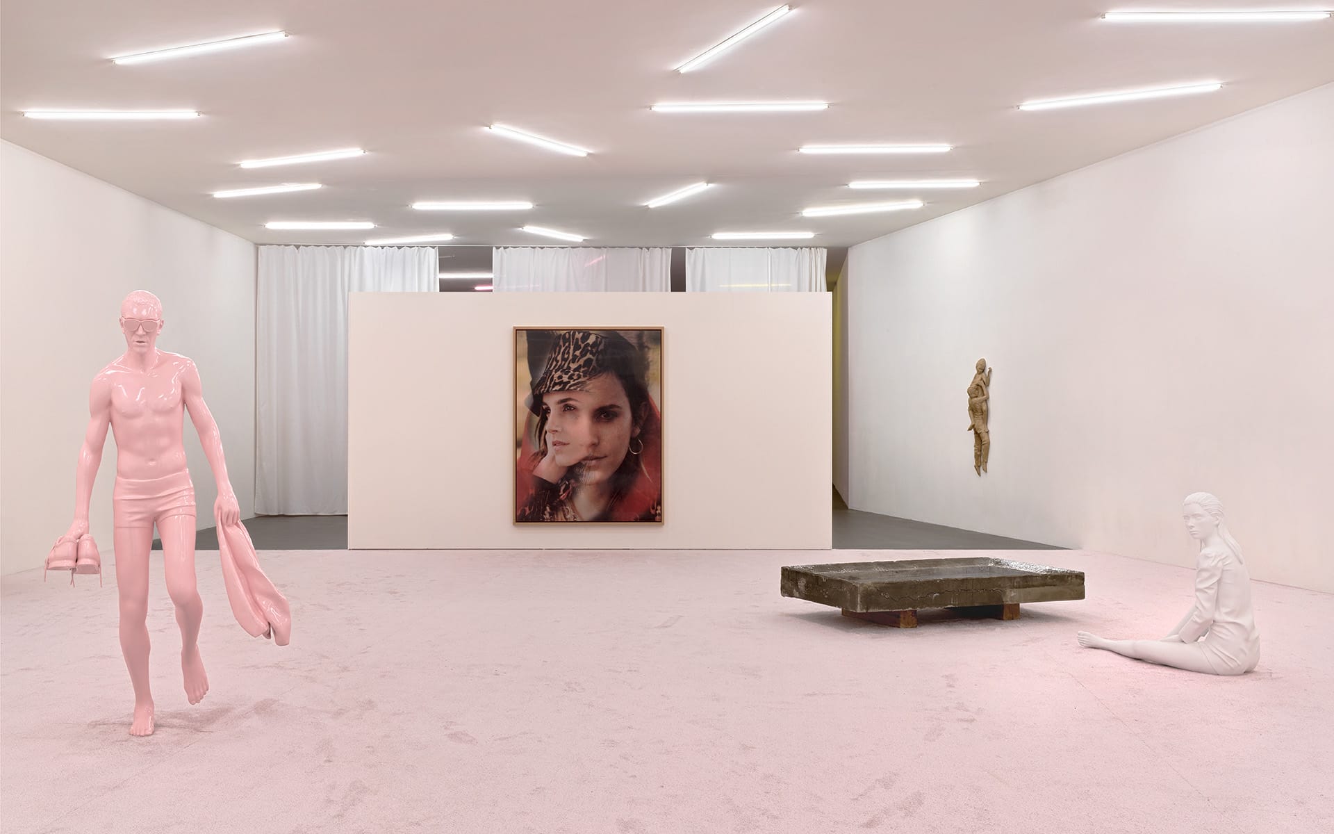 Art gallery with sculptures and a portrait on display, pink statue walking, seated figure, and gold relief on white walls.