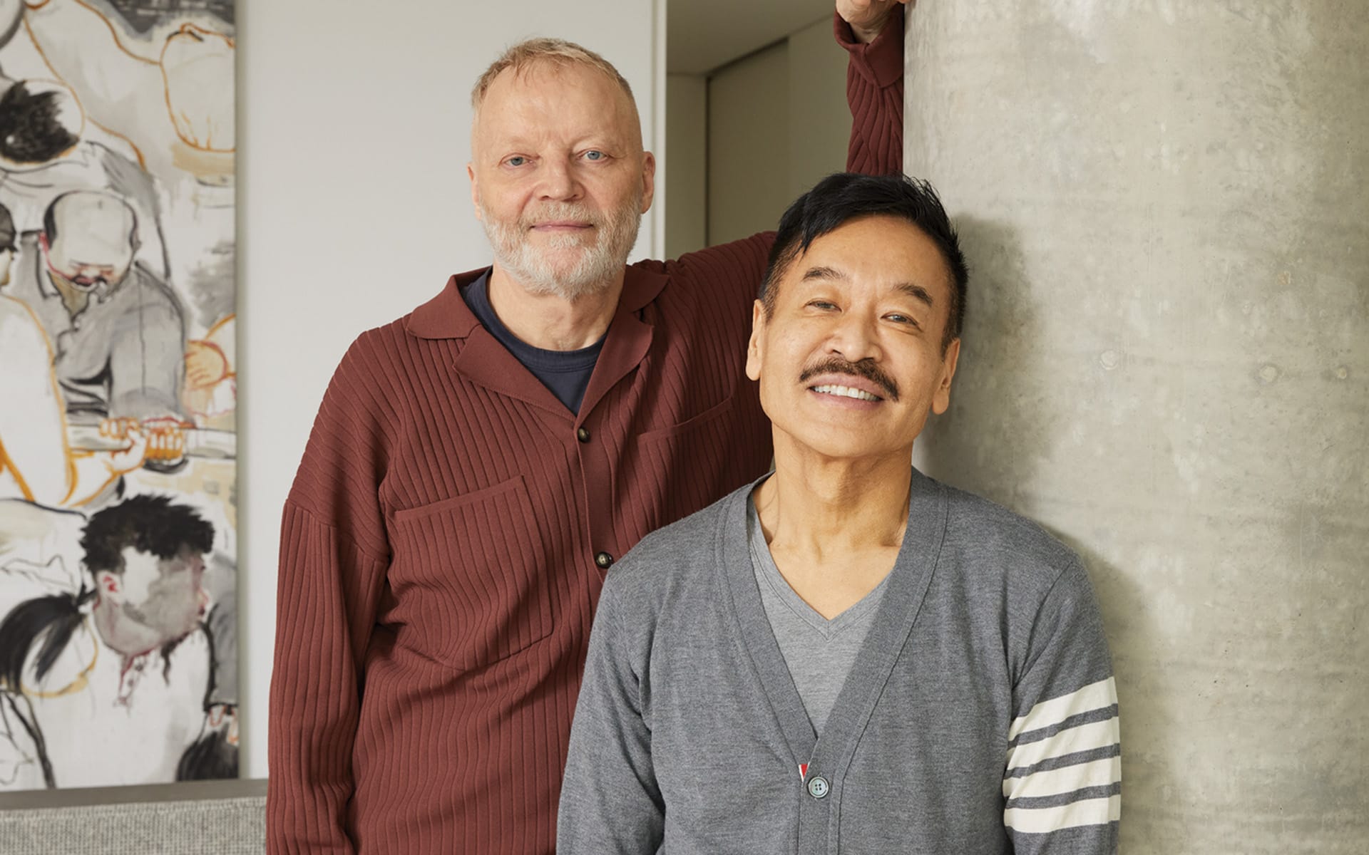 Two smiling men standing indoors near an artwork and a concrete pillar.