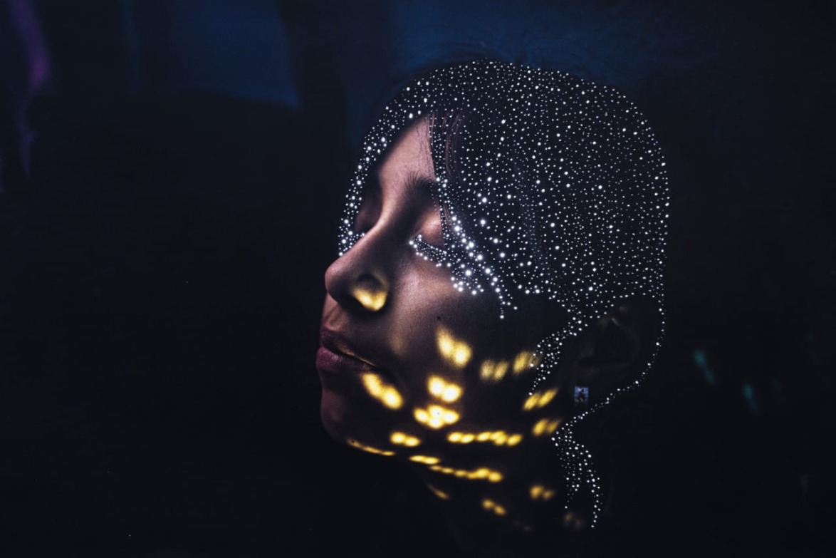 Person with illuminated dots and patterns on face in a dark setting, eyes closed, creating a mystical atmosphere.