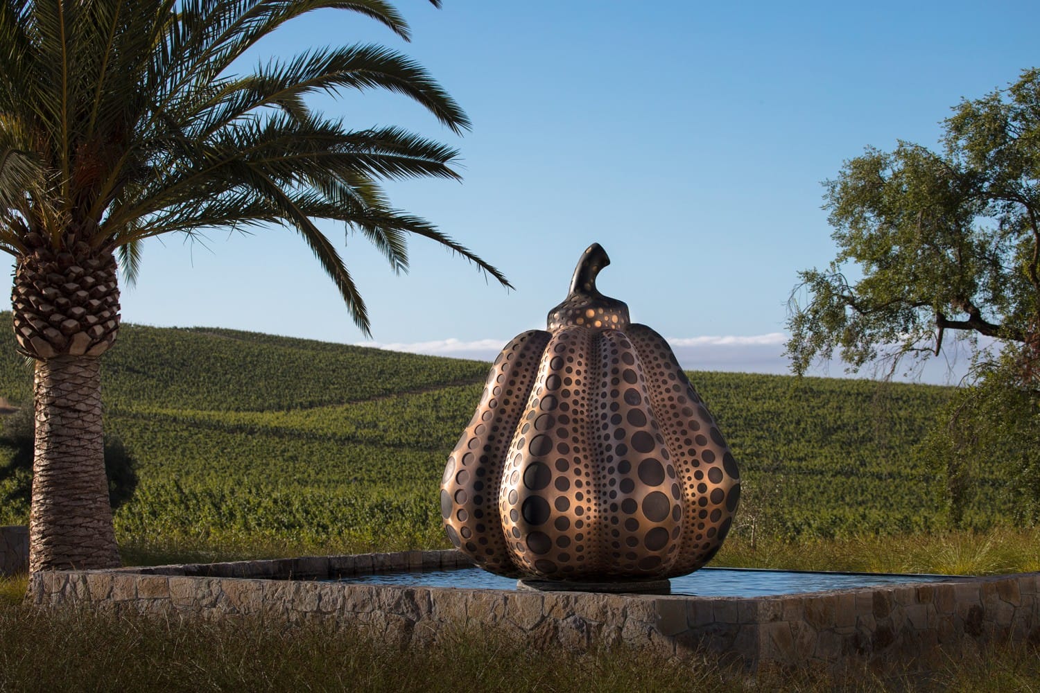 Large metallic pumpkin sculpture with circular patterns, set in a field with a palm tree and rolling hills in the background.
