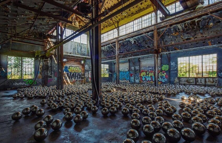 Abandoned warehouse with metal structures and hundreds of metallic spheres on the floor, surrounded by graffiti-covered walls.