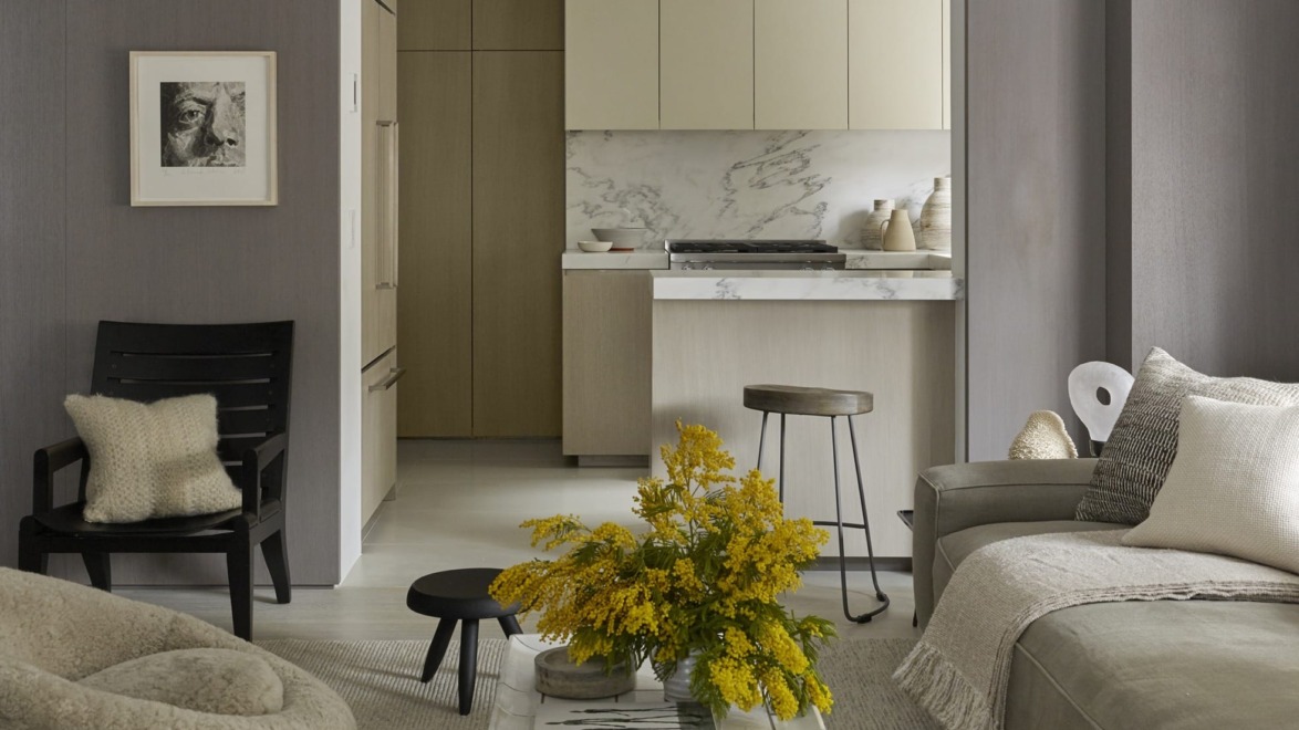 Modern living room with beige sofa, yellow flowers on table, and view of minimalist kitchen in the background.