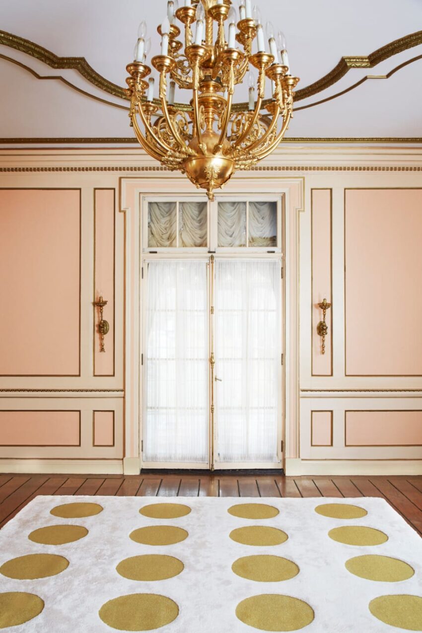 Elegant room with a large gold chandelier, ornate cream and gold walls, and a polka-dot patterned carpet on wooden floor.