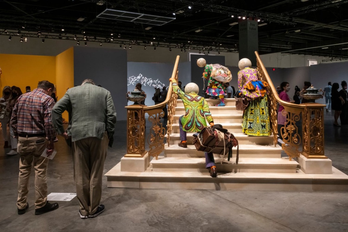 People observing vibrant, colorful art figures walking up ornate stairs inside an exhibition hall.