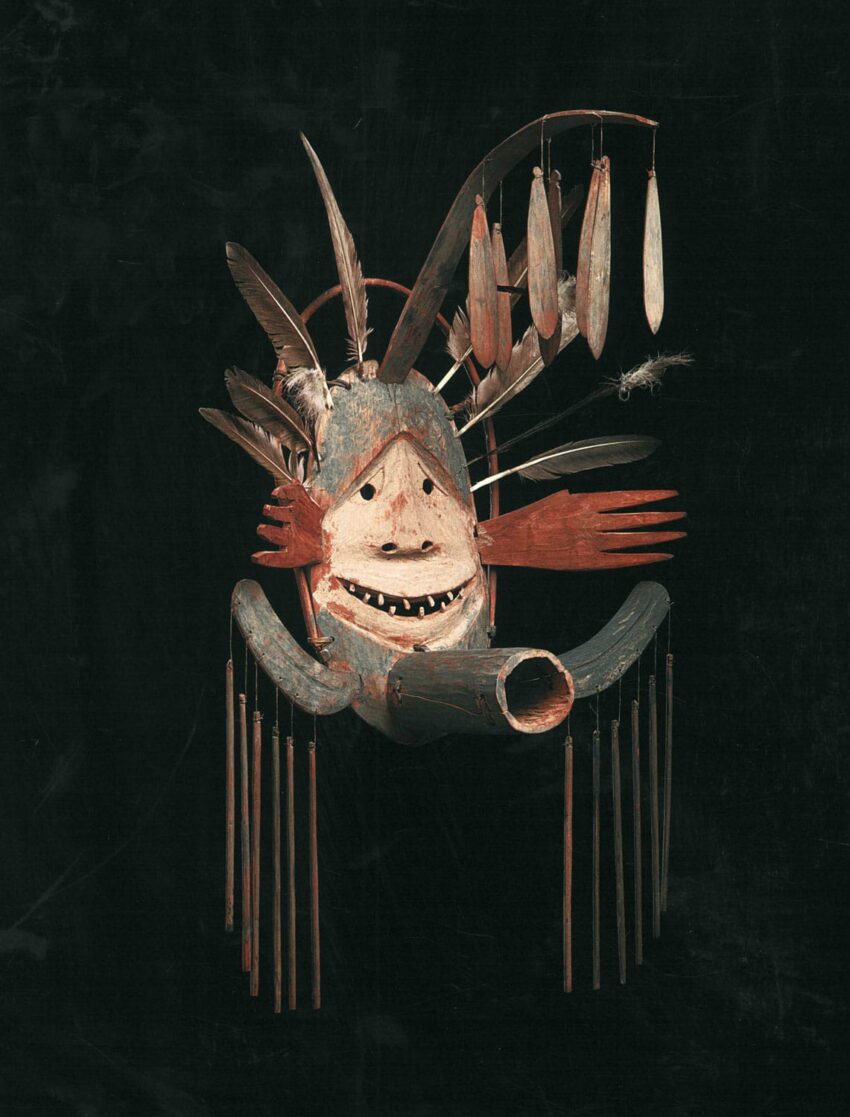 Traditional wooden mask with feathers, depicting a face with a wide smile and red accents on a dark background.