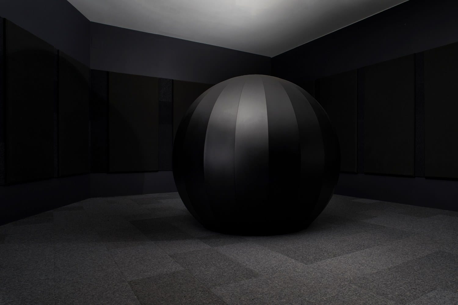 A large black sphere in a dark room with gray carpeted floor and black walls.