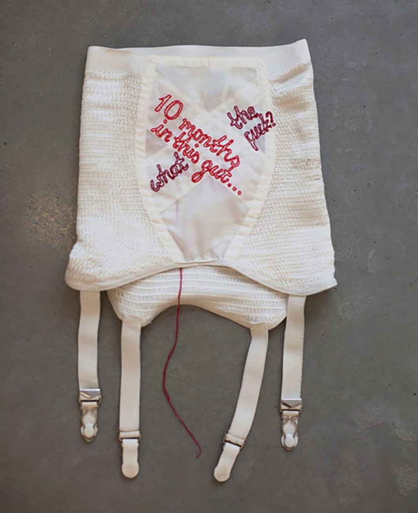 White vintage corset with red embroidered text asking "10 months in this gut... The result?" displayed on a gray background.
