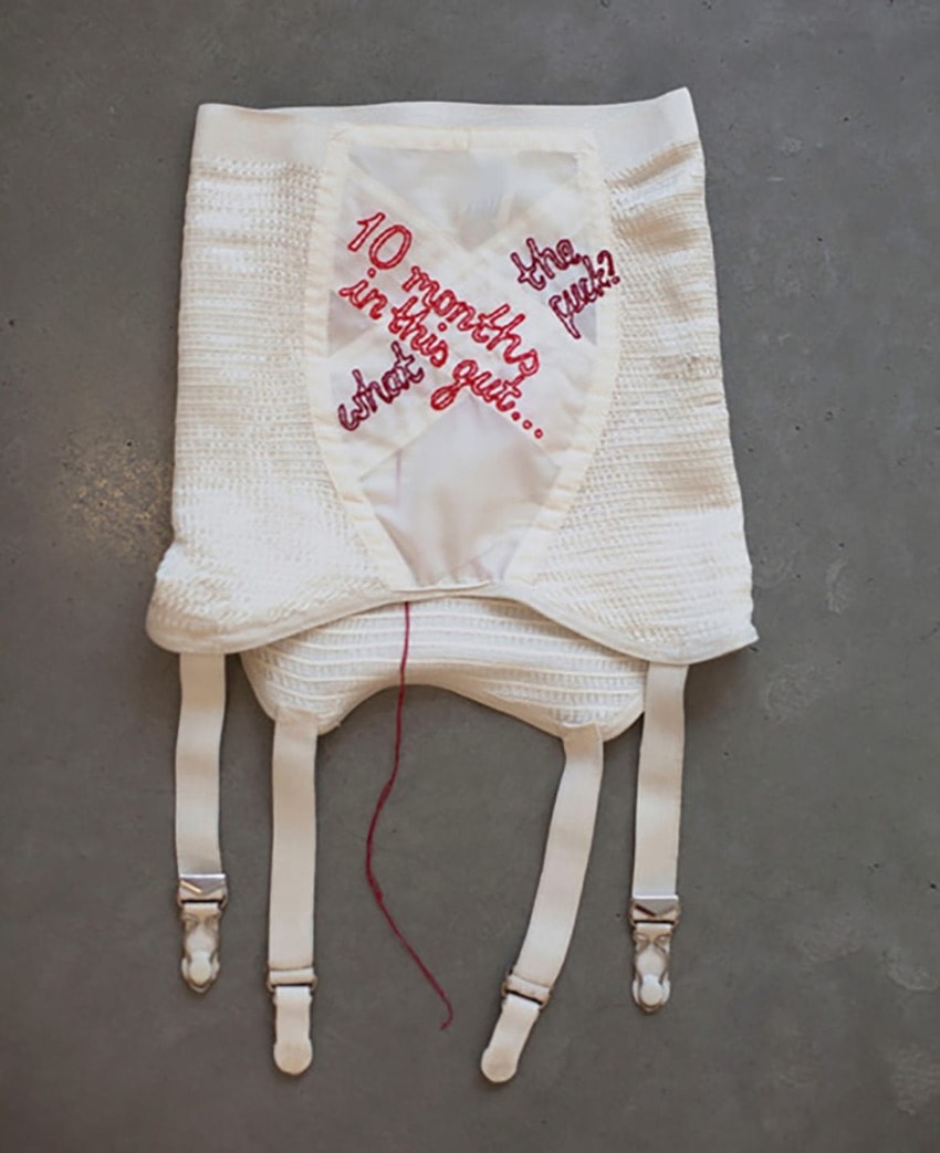 White vintage corset with red embroidered text asking "10 months in this gut... The result?" displayed on a gray background.