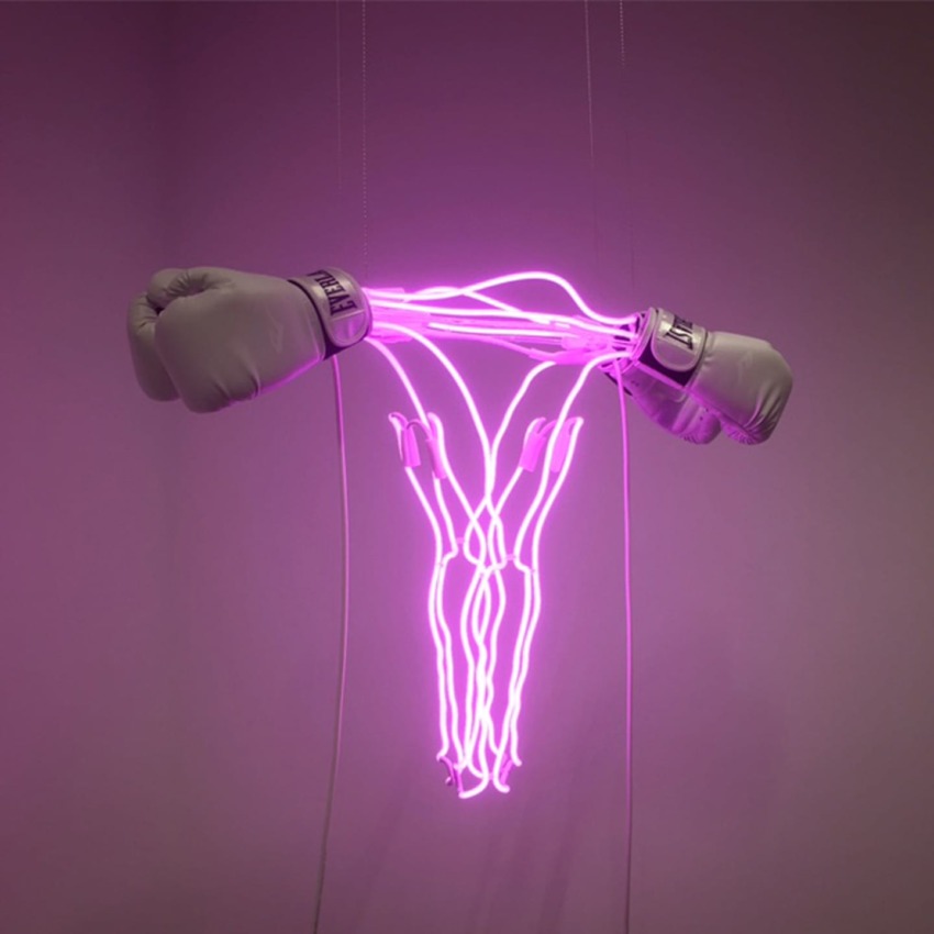 Glowing pink neon lights shaped like a uterus with boxing gloves on each side hanging against a plain wall.