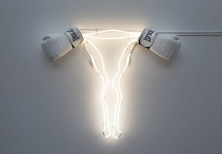 Uterus-shaped neon light with boxing gloves attached at ends, mounted on a plain wall.