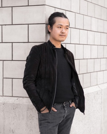 Person in black jacket leans against a brick wall, looking thoughtful and relaxed.