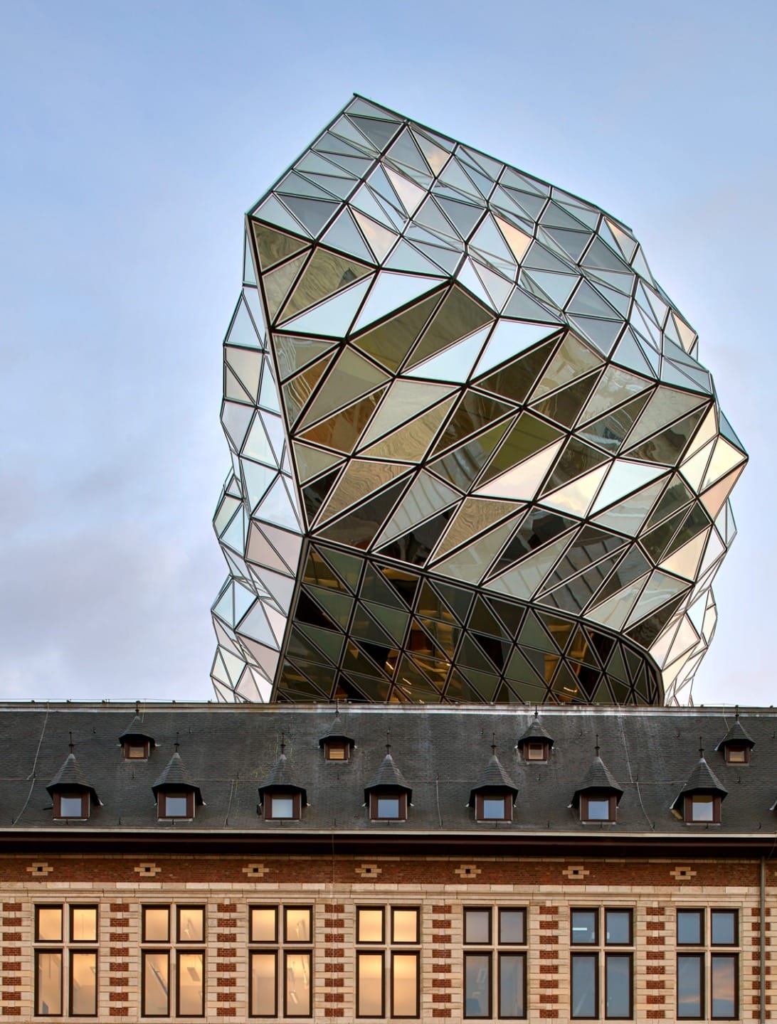 Modern glass building with geometric design on top of a historic brick structure against a clear sky background.