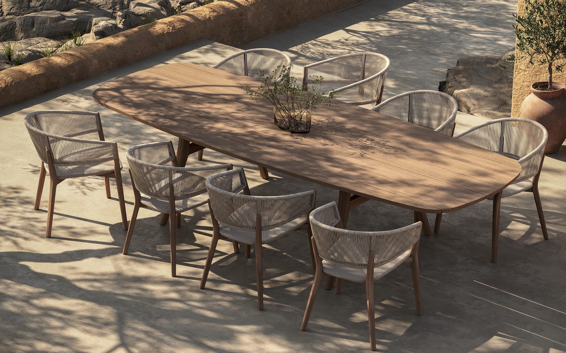 Outdoor dining set with a large wooden table and ten cushioned chairs on a sunlit patio, featuring a centerpiece plant.