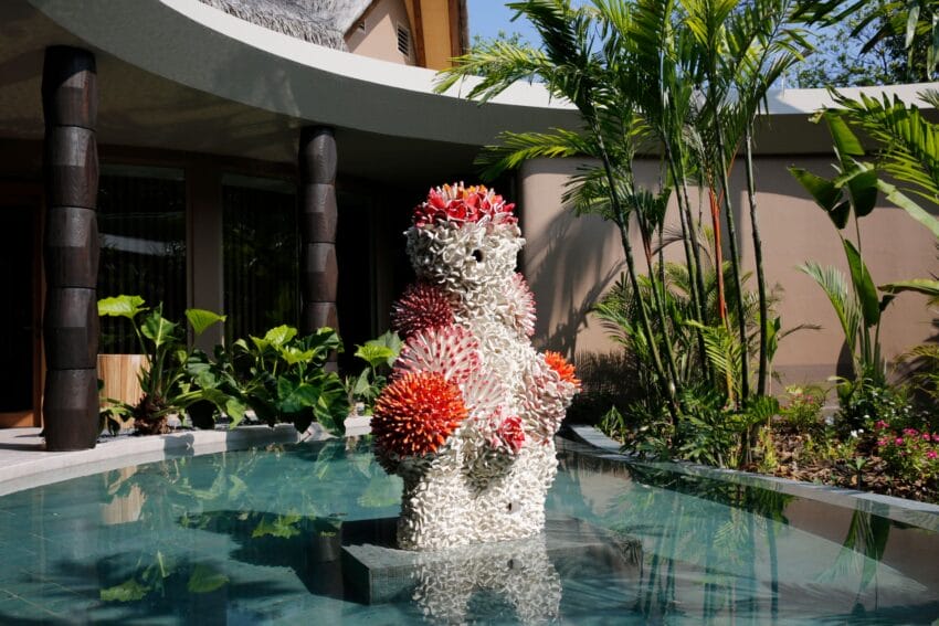 Modern sculpture with vibrant colors standing in a tranquil outdoor courtyard surrounded by lush green plants and water.