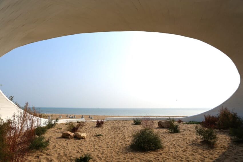 Architectural structure framing a serene beach view with sandy shore, sparse vegetation, and calm ocean under a clear sky.