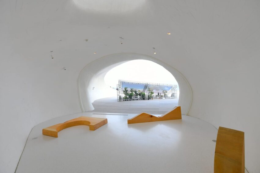 Minimalist interior space with curved white walls, wooden seating, and a window view of an outdoor area with greenery.
