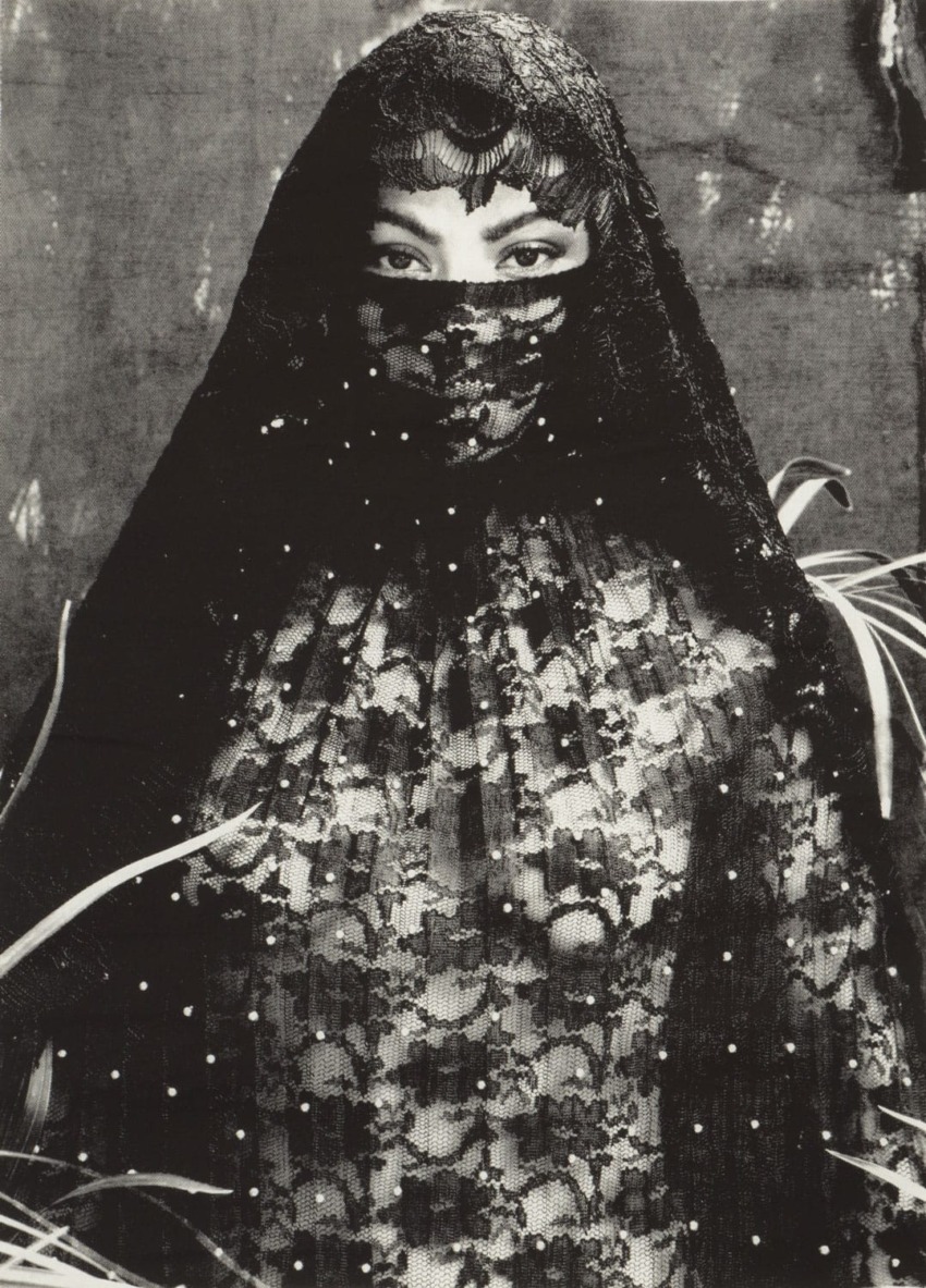 Person in lace veil and attire standing amidst foliage.