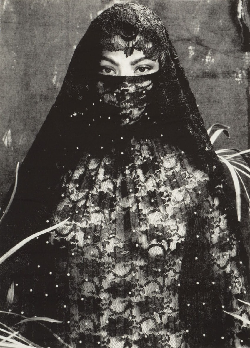Person in lace veil and attire standing amidst foliage.