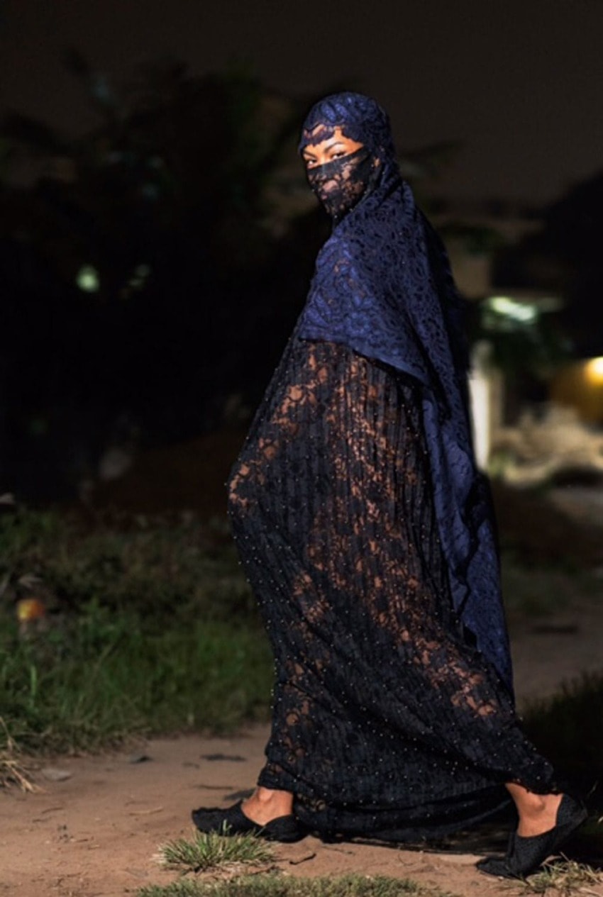 Person in intricate dark veil walks outdoors at night, surrounded by blurred greenery and dim lighting.