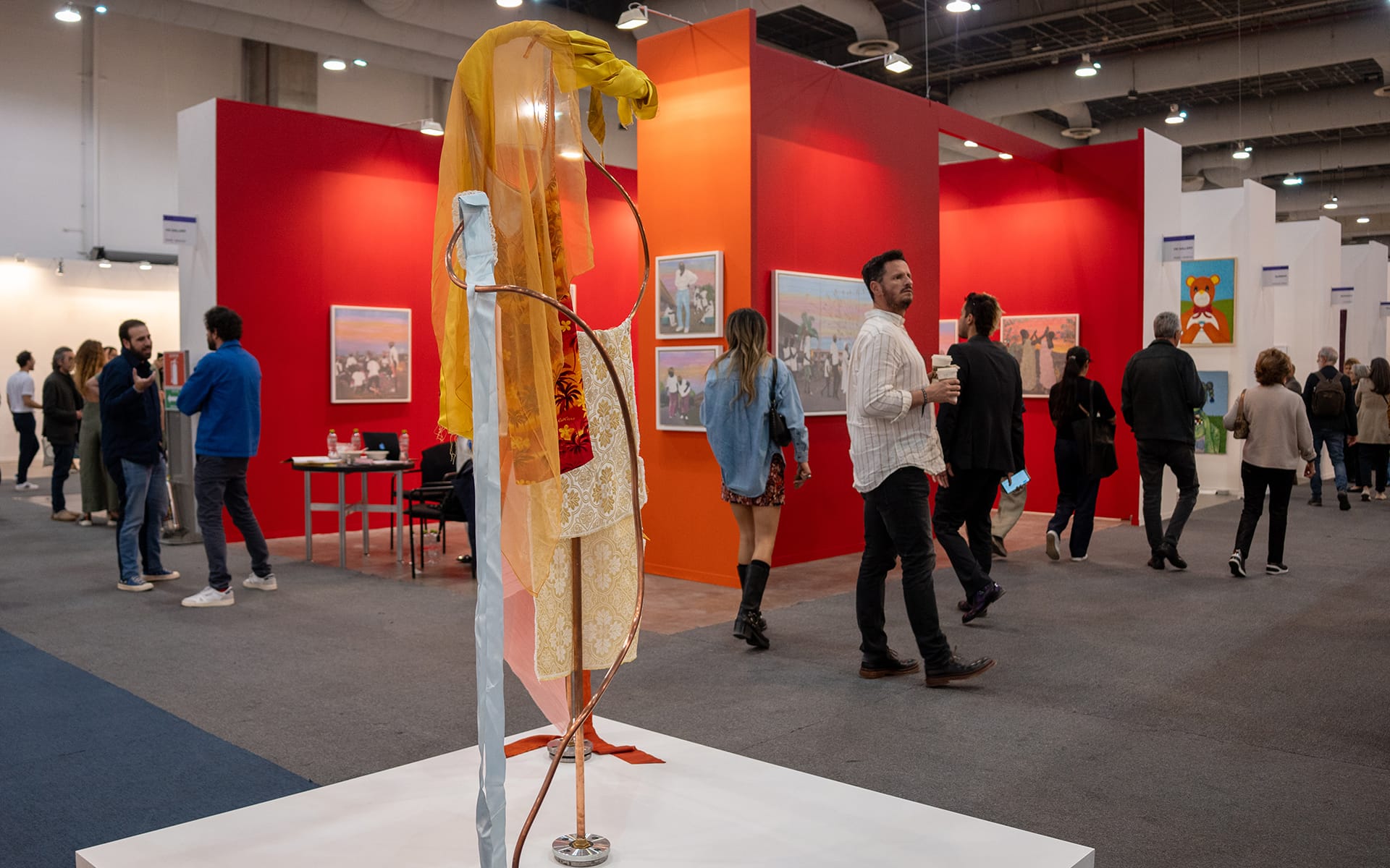 Art exhibition with colorful sculptures and paintings, people walking and observing artworks in a large, well-lit gallery space.