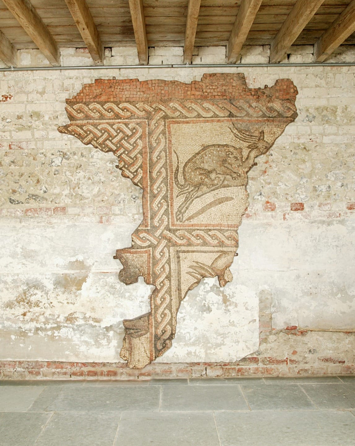 Ancient Roman mosaic with intricate patterns and a deer design, displayed on a worn brick wall under a wooden ceiling.