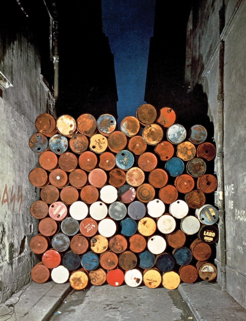 Wall of stacked rusty and painted barrels in a narrow alleyway under a dark blue sky, flanked by graffiti-marked walls.