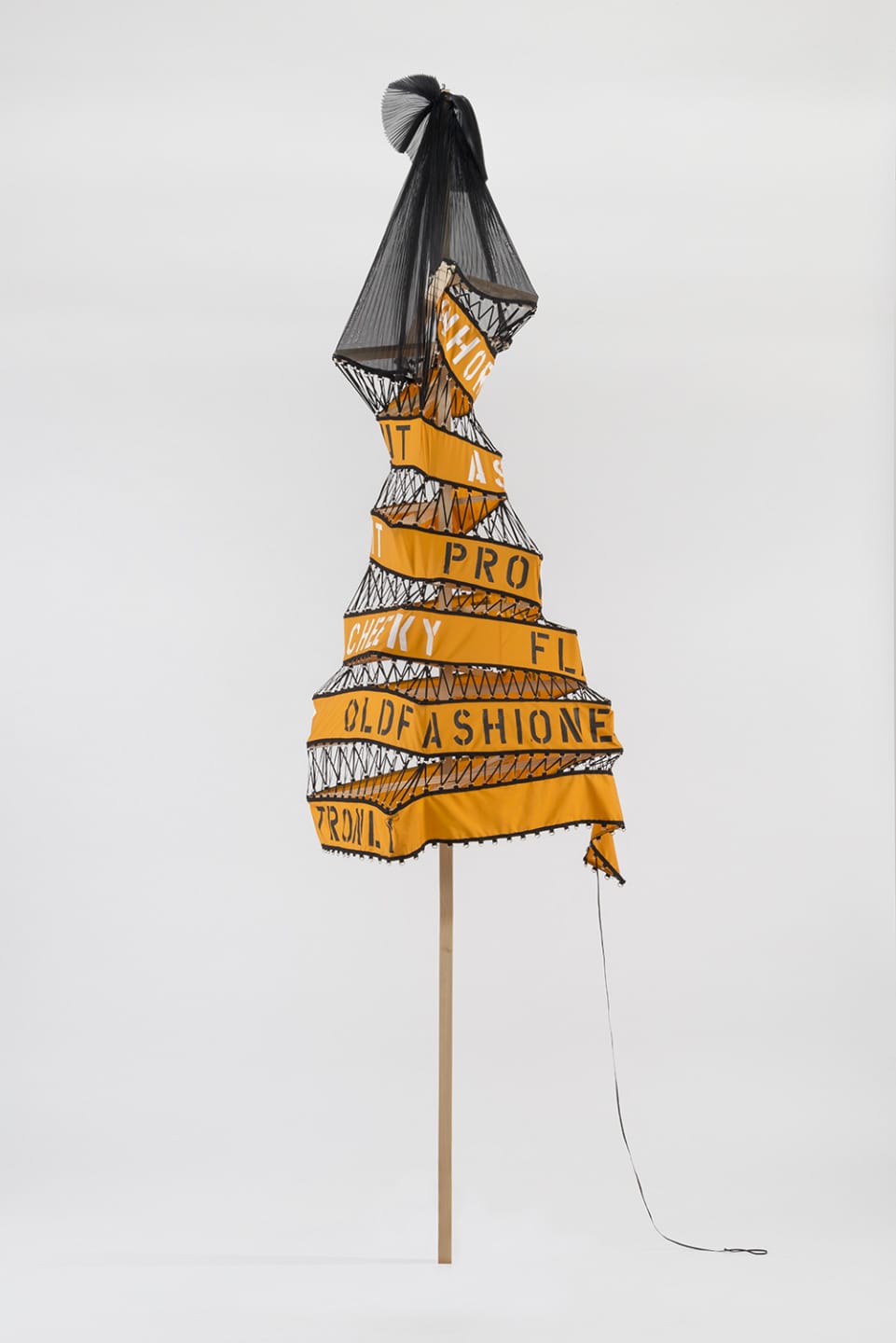 Artistic installation of a dress-like sculpture with yellow and black text, mounted on a wooden pole against a white background.