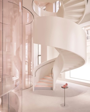 Spiral staircase in modern, minimalist interior with glass walls and a pair of red high heels displayed on a small stand.