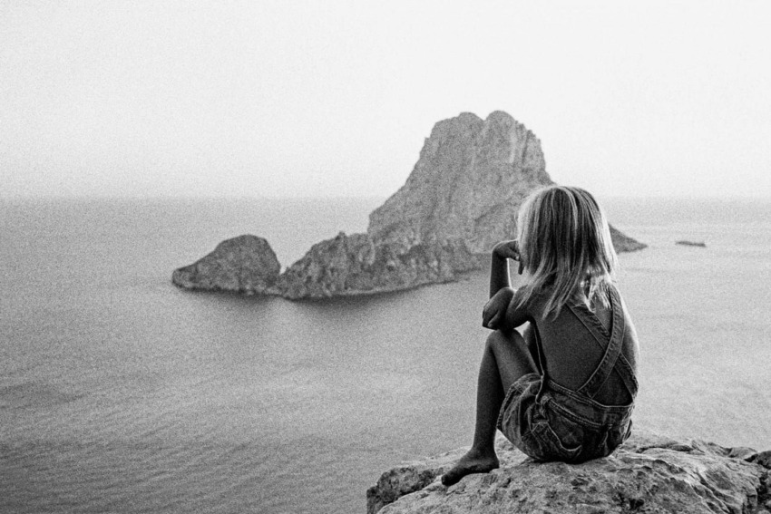 Child sitting on a rock overlooking the sea with distant rocky islands in black and white.