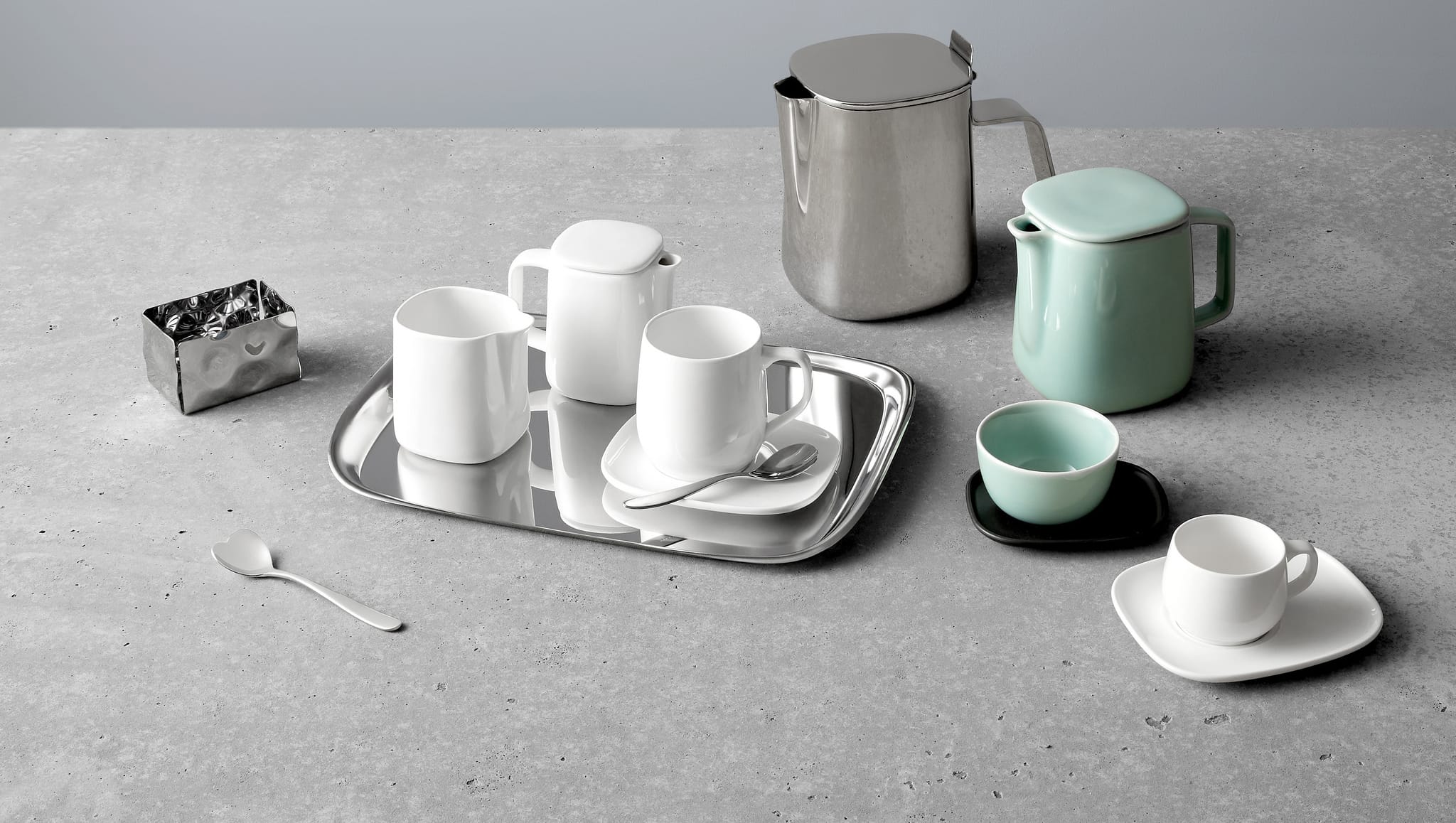 Minimalist tea set on a gray countertop with cups, saucers, teapots, a tray, and a spoon, featuring white and mint colors.