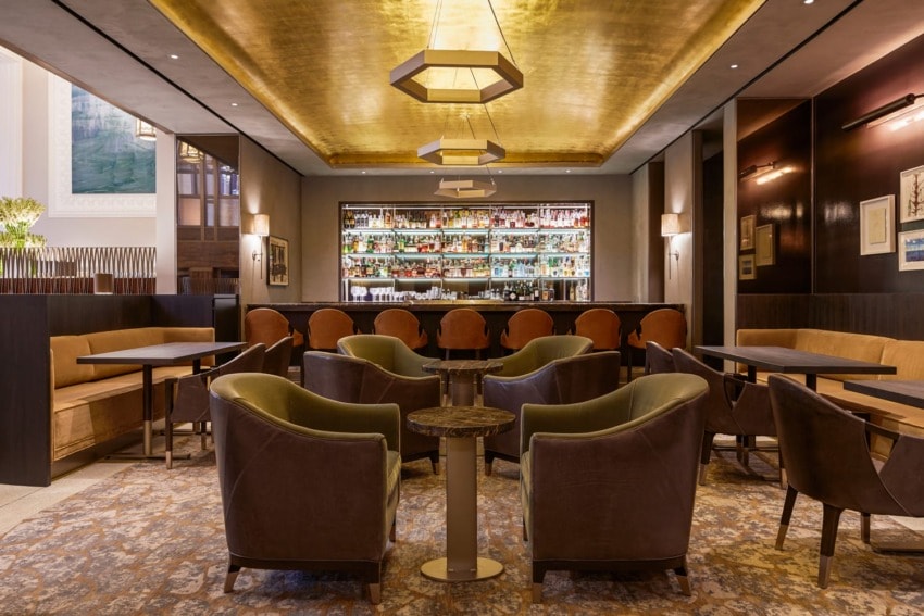 Elegant hotel bar with modern decor, green and brown chairs, round tables, and a well-stocked liquor shelf in the background