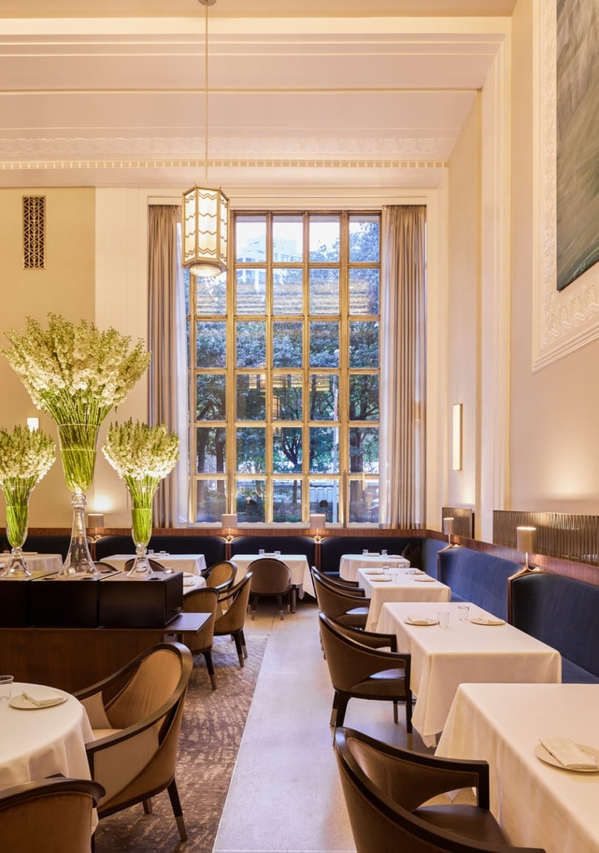 Elegant restaurant interior with large windows, tall flower arrangements, and neatly set tables with white tablecloths.