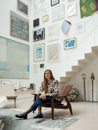 Woman sitting in a chair in an art-filled room with framed artwork on the walls and sculptures nearby.