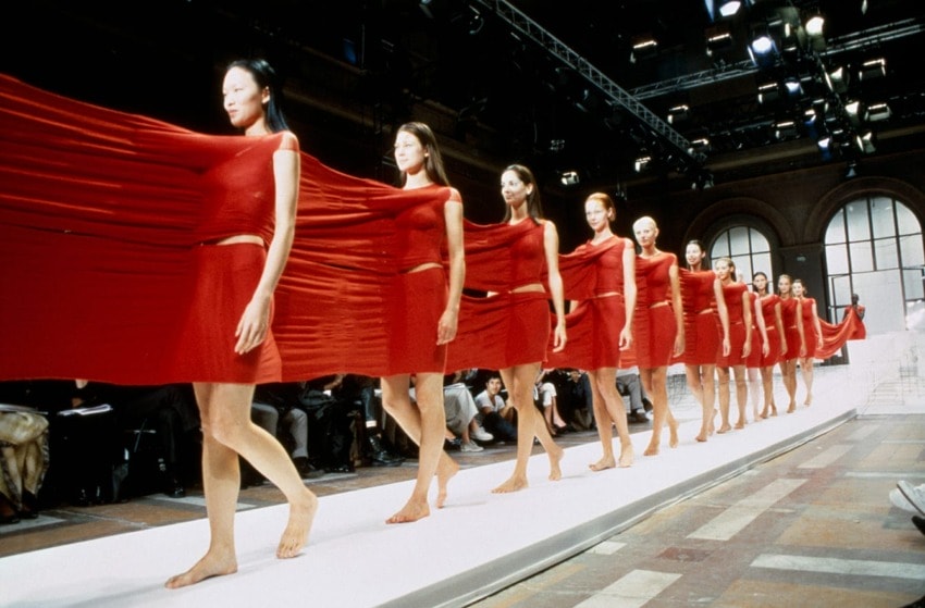 Models walk in a fashion show wearing interconnected red outfits, each part of a continuous fabric drape.