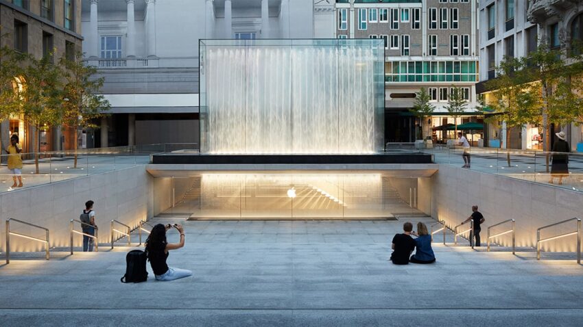 Underground entrance to an Apple Store with a glass waterfall feature, surrounded by people sitting on steps.