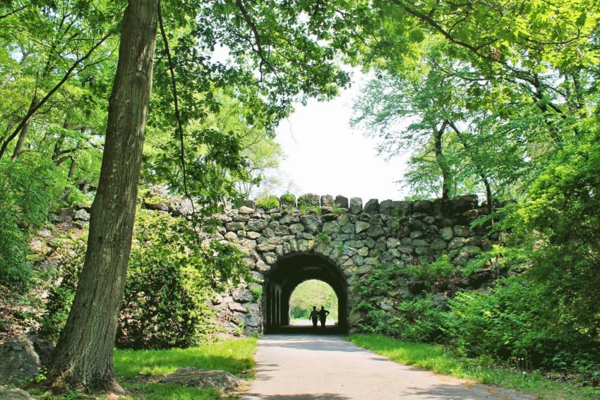 Stone arch bridge in a lush park with two people walking on the path underneath.