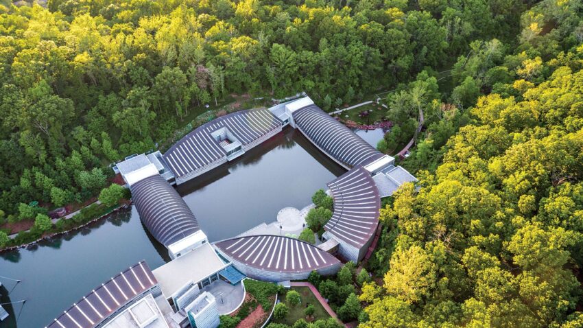 Aerial view of a museum with curved roofs surrounded by lush green forest and a tranquil pond.