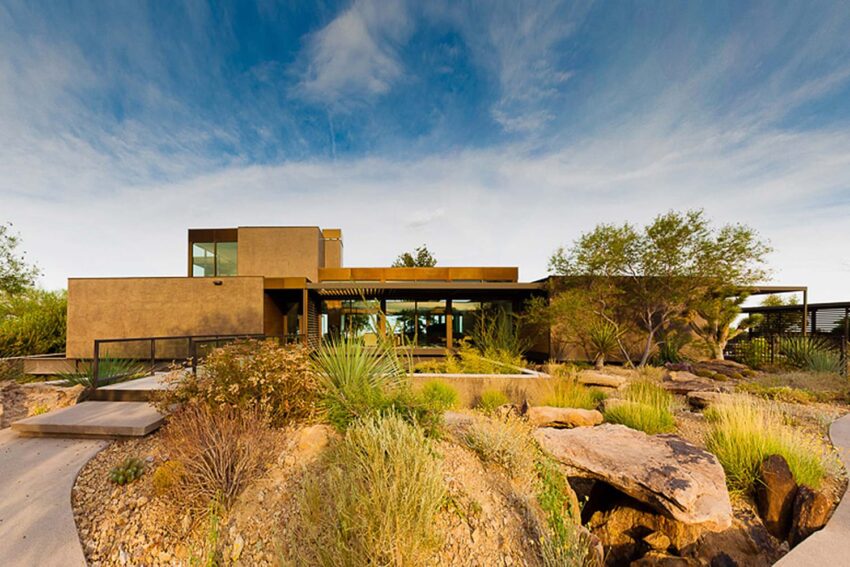 Modern house with large glass windows surrounded by desert landscape, featuring rocks and native plants under a blue sky.
