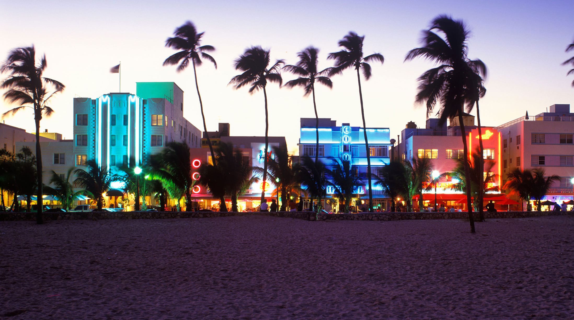 Art Deco buildings with neon lights on Ocean Drive in Miami Beach, palms swaying under a dusky sky.